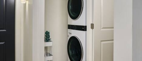 A laundry room with two stacked front-loading washing machines, a white shelving unit with laundry supplies, and a dark door.