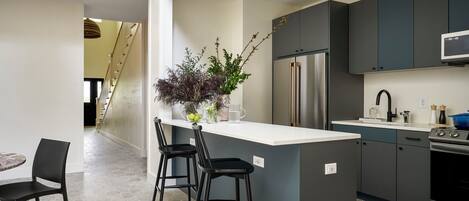 A modern kitchen with a central island, bar stools, a stainless steel refrigerator, and a microwave.