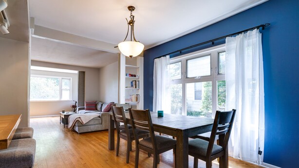 Un comedor con mesa y sillas de madera, una estantería y un ventanal con cortinas.