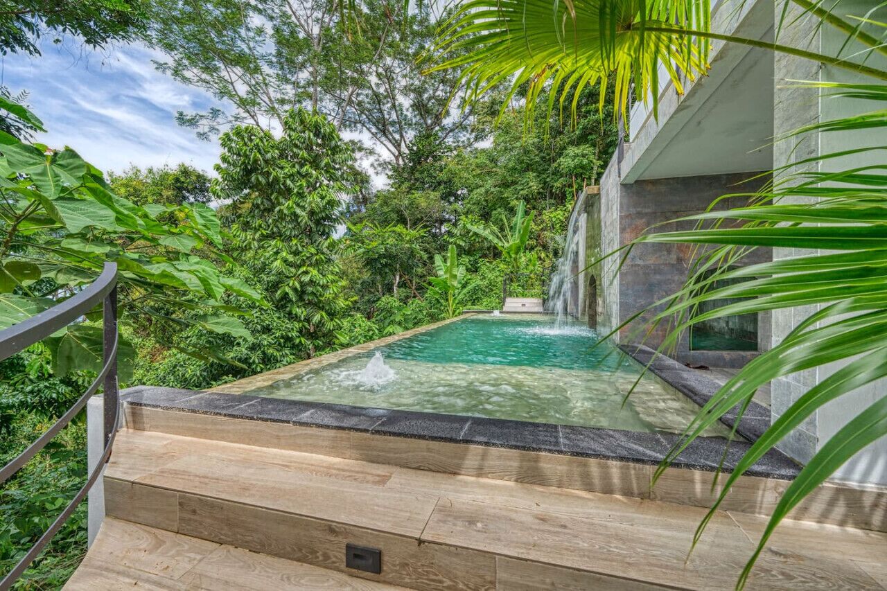 A swimming pool with a waterfall feature, surrounded by lush greenery and a stone deck.