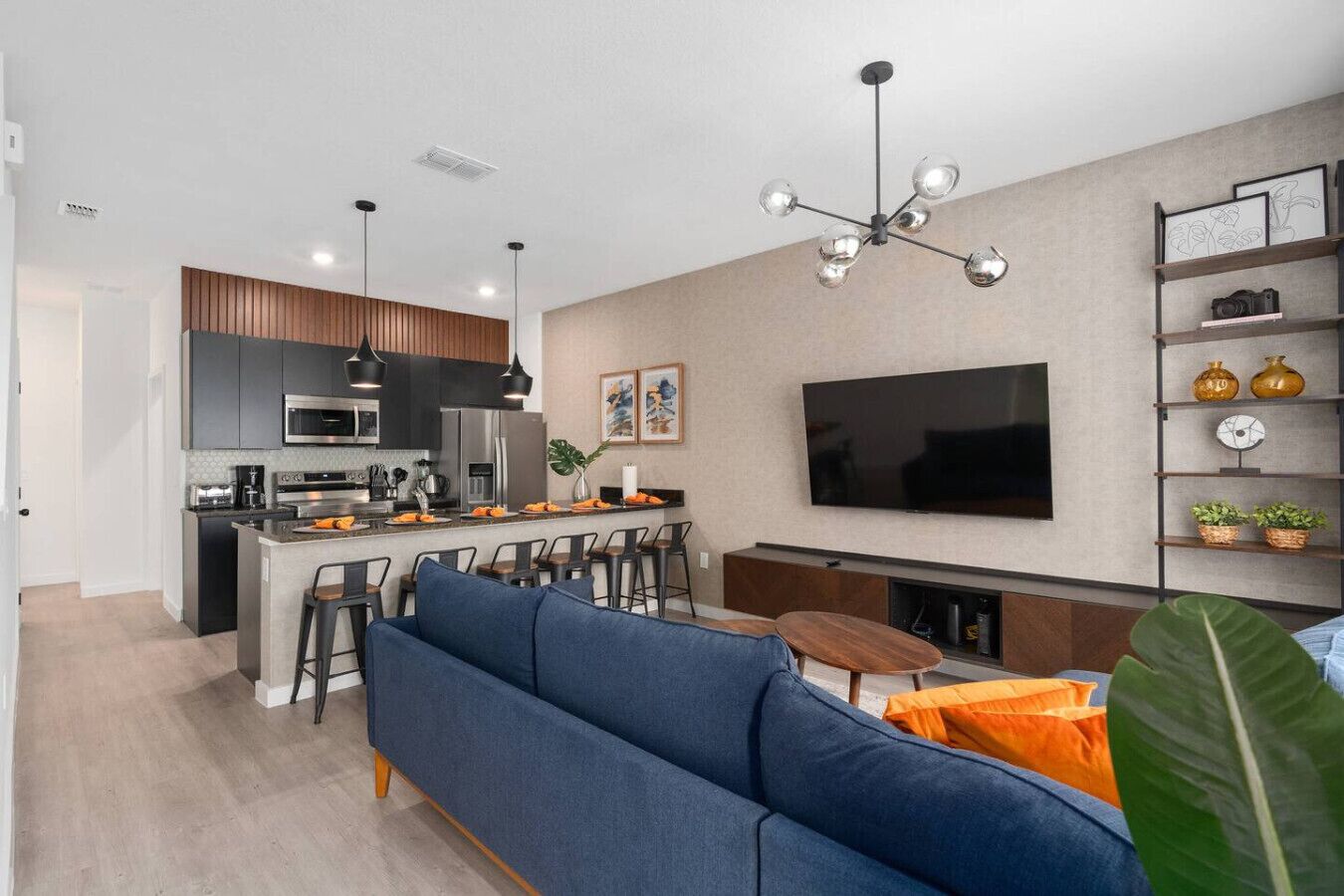 A modern kitchen with a central island, a dining area with a sofa and chairs, a flat-screen TV mounted on the wall, and a bookshelf with decorative items.