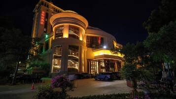A multi-story hotel with a curved facade, illuminated entrance, and a parking area with a car.