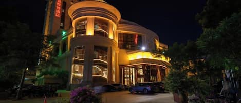 A multi-story hotel with a curved facade, illuminated entrance, and a parking area with a car.