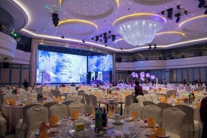 Un salón de banquetes con mesas redondas, sillas y un escenario para presentaciones.