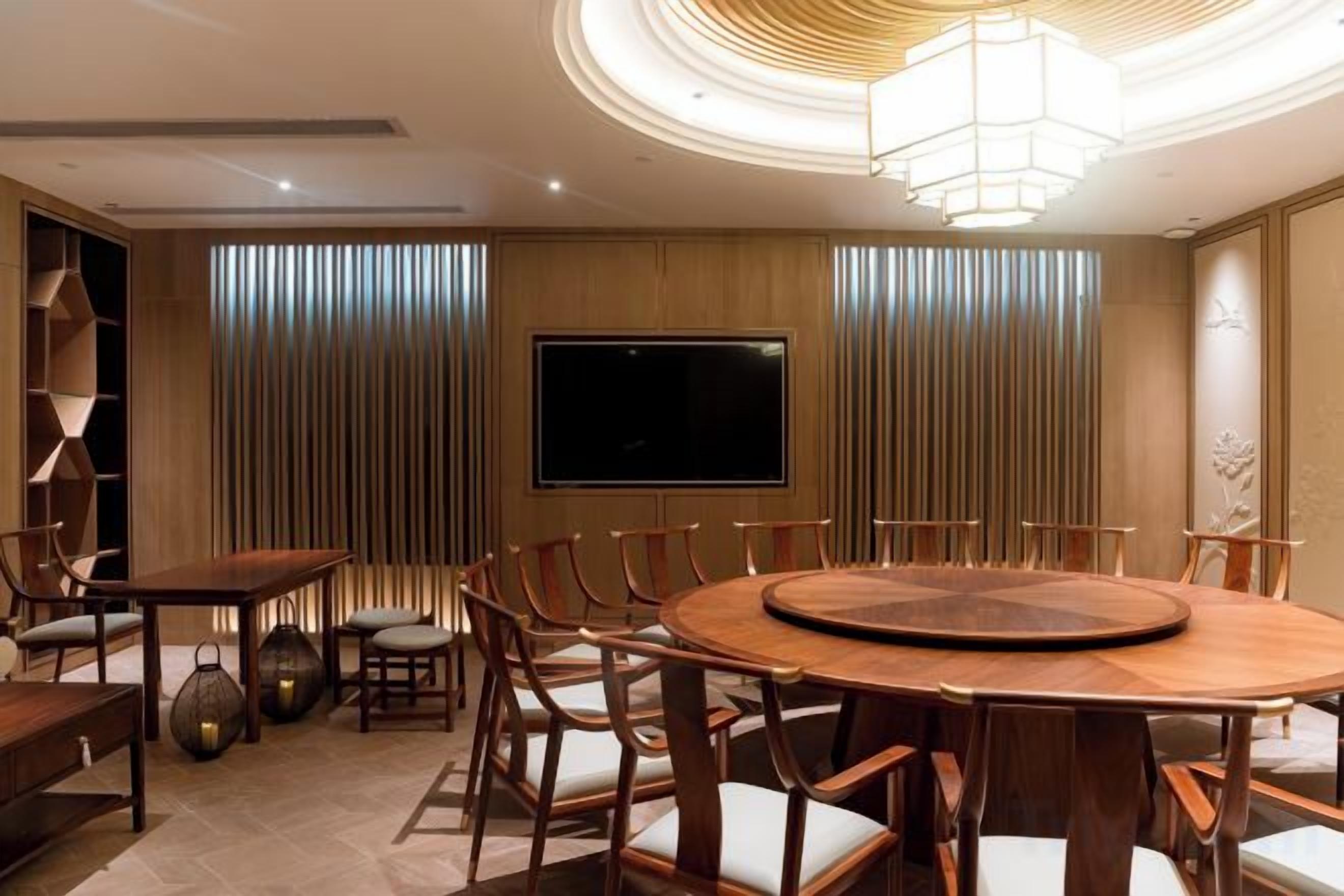 A modern dining area with a round wooden table surrounded by chairs, a television mounted on the wall, and a decorative ceiling light.