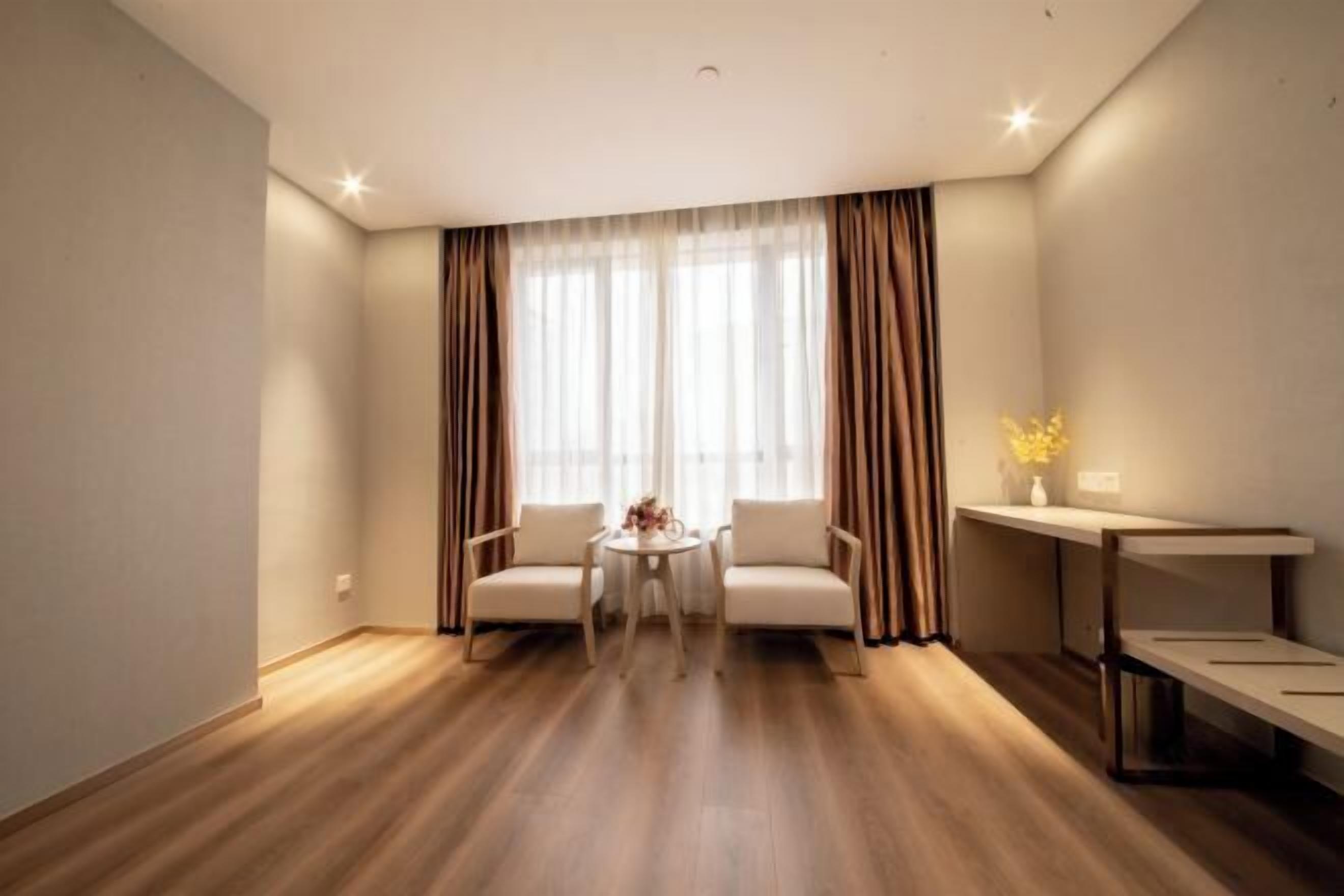 A modern hotel room with a wooden floor, two white armchairs, a small round table, a desk with a chair, and a large window with sheer curtains.