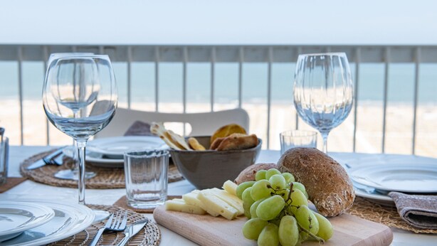 Ein Tisch ist für eine Mahlzeit gedeckt, mit Tellern, Besteck, Gläsern und einer Schüssel mit Brot und Trauben, mit Blick auf einen Strand.