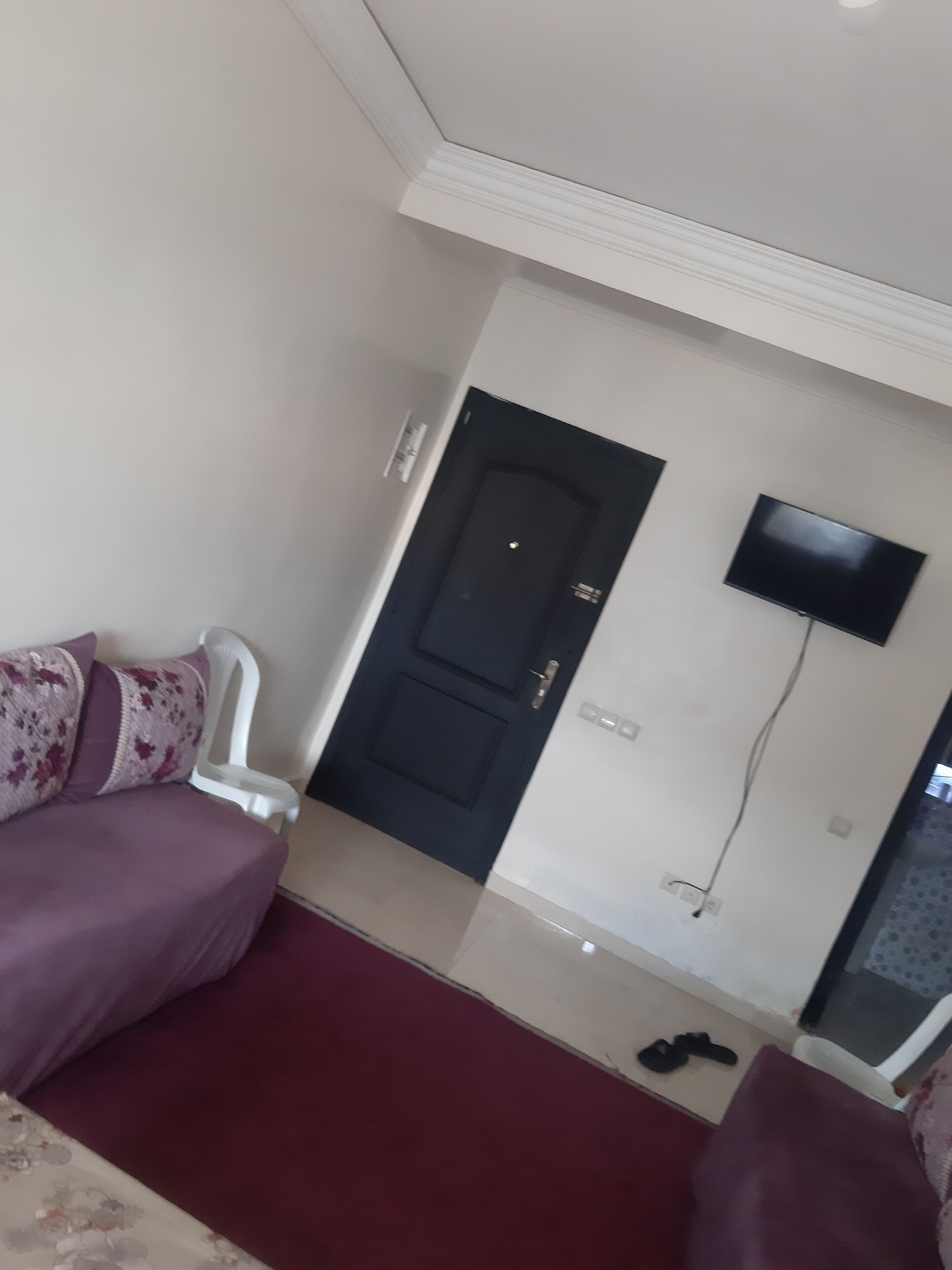 Un salon avec un canapé, un téléviseur fixé au mur, une porte et une paire de chaussures posées sur le sol.