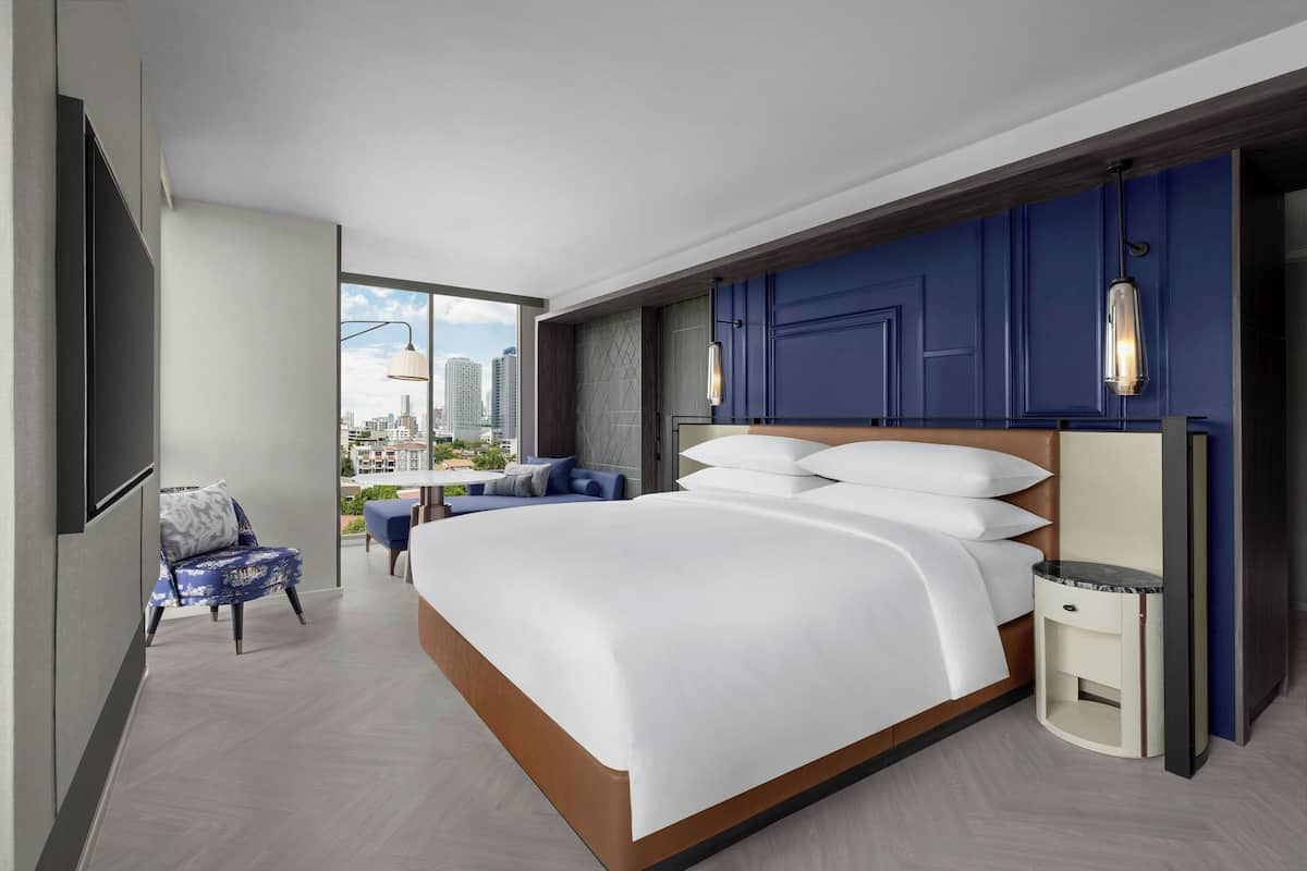 A modern hotel room with a large bed, a seating area with a chair and table, and a city view through a large window.
