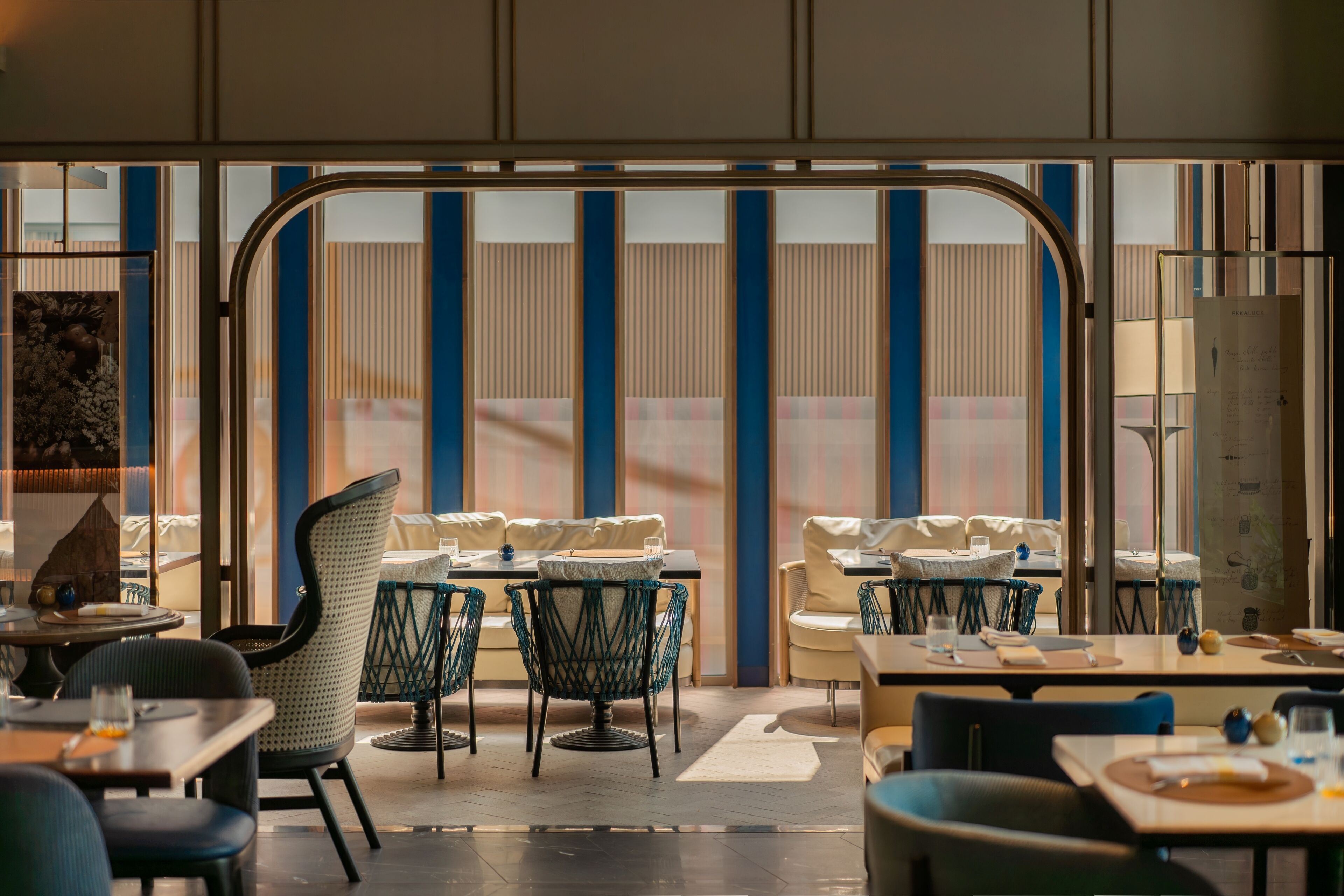 A modern restaurant with a view through large windows, featuring a mix of seating arrangements including tables and chairs.