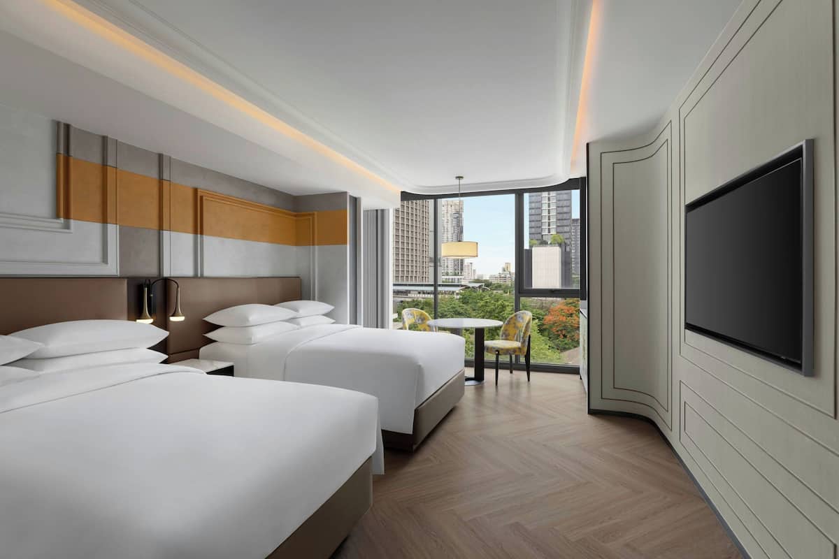 A hotel room with two beds, a TV, a balcony with a view of the city, and a small table with chairs.