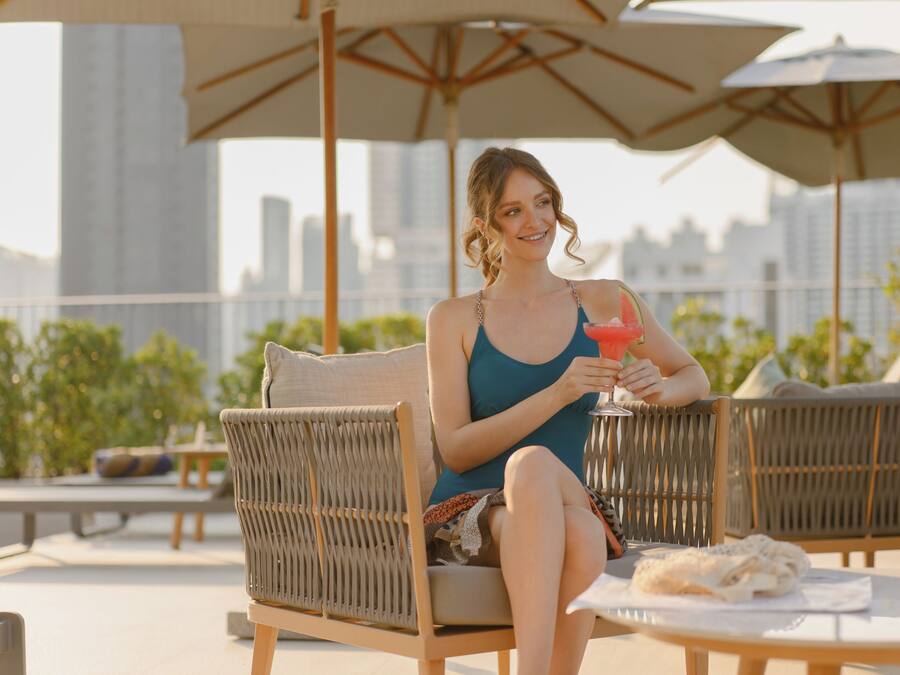Eine Person sitzt auf einem Korbstuhl, einen Drink haltend, mit Blick auf eine Stadtlandschaft.