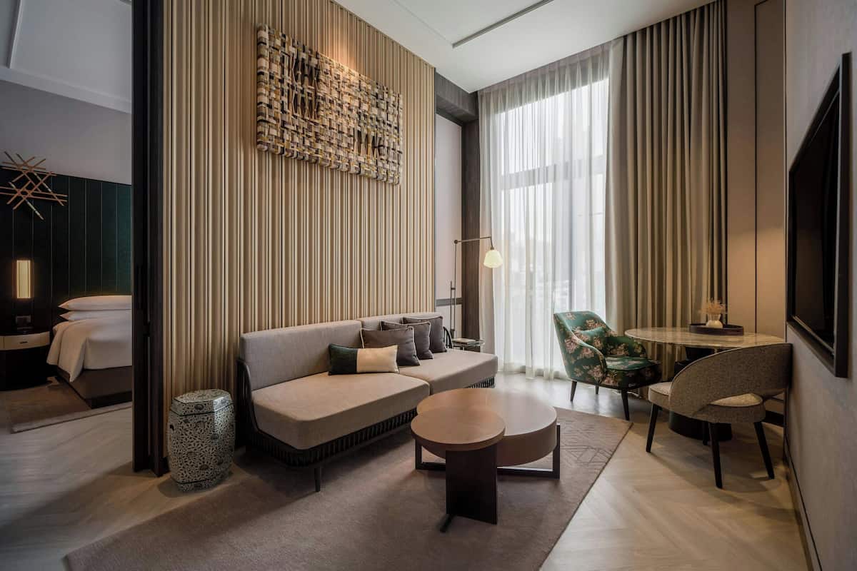 A modern hotel room with a sectional sofa, a round coffee table, a small side table, a decorative wall piece, and a large window with sheer curtains.