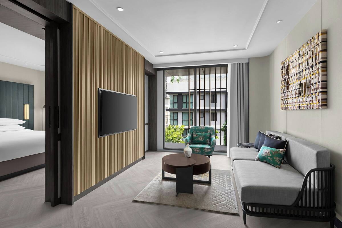 A modern hotel room with a bed, a sofa, a round coffee table, and a TV mounted on the wall.