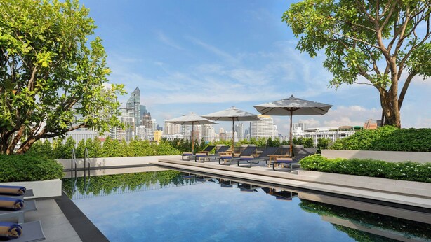Piscina en la azotea con sillones de descanso y sombrillas, rodeada de vegetación y vistas al horizonte urbano.