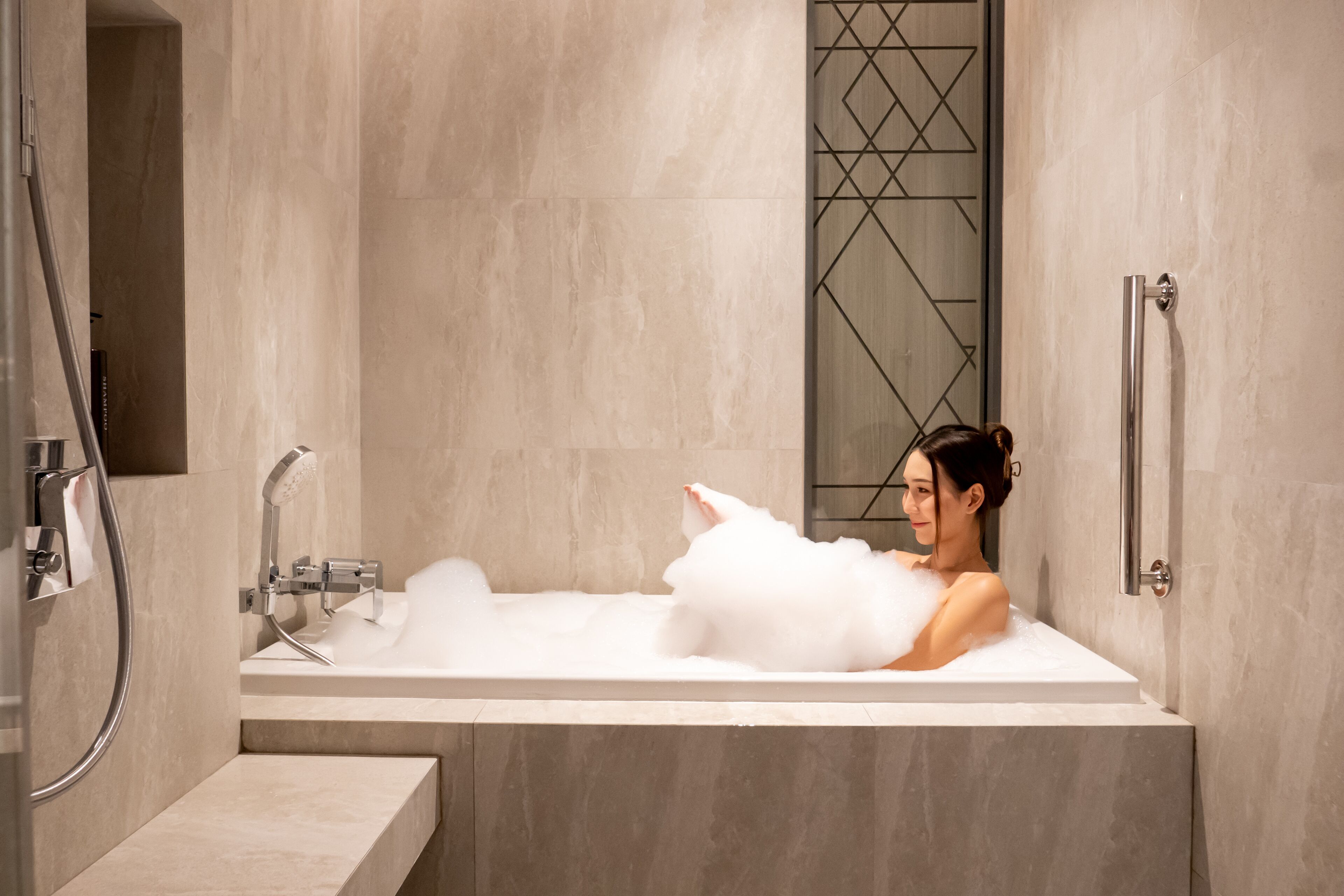 Una mujer en una bañera con un gato blanco, rodeada de un moderno baño con un separador de vidrio geométrico y un pasamanos.