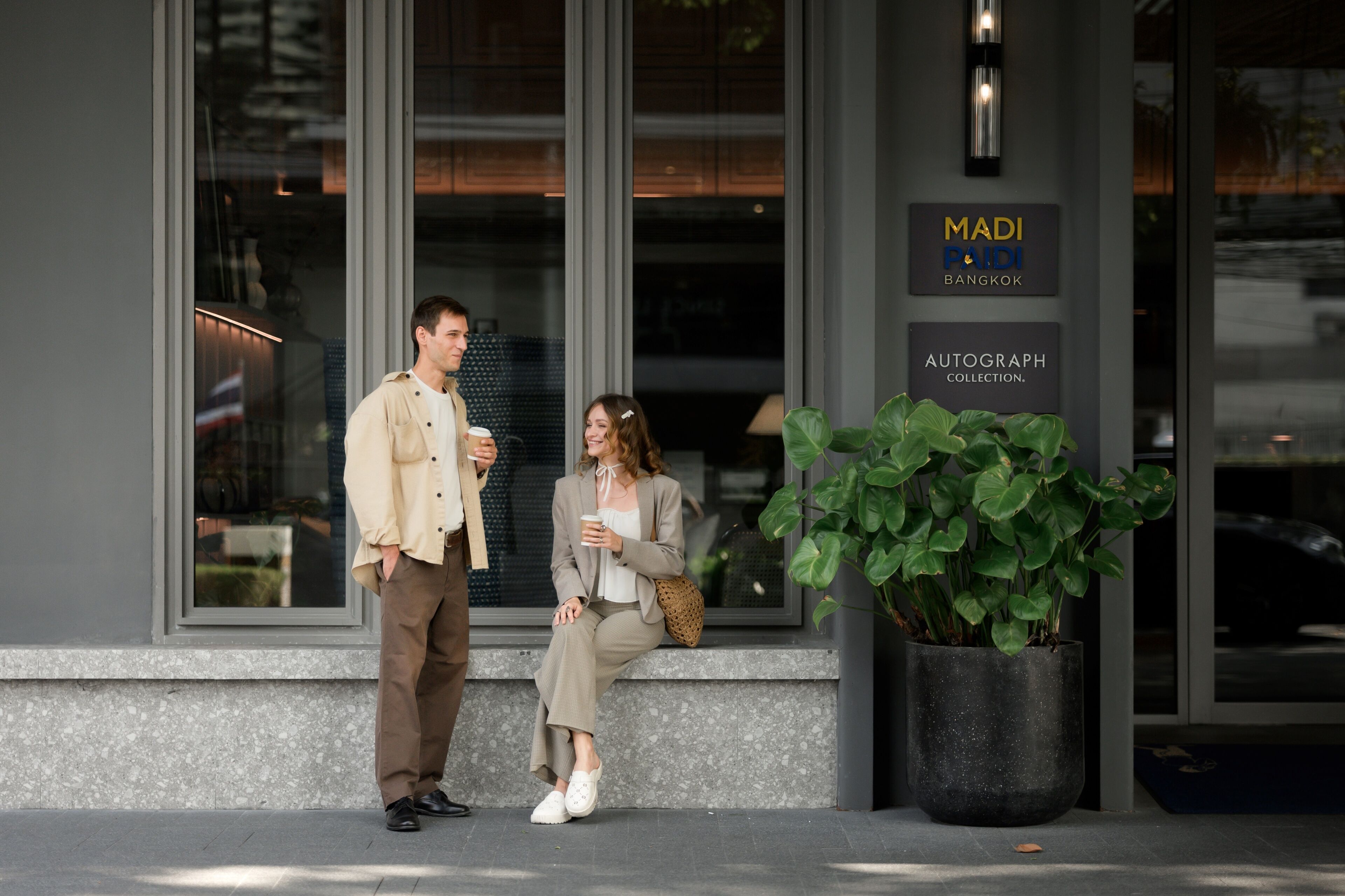 Two people standing outside a building with a sign that reads 'MADI POOI BANGKOK AUTOGRAPH COLLECTION.'