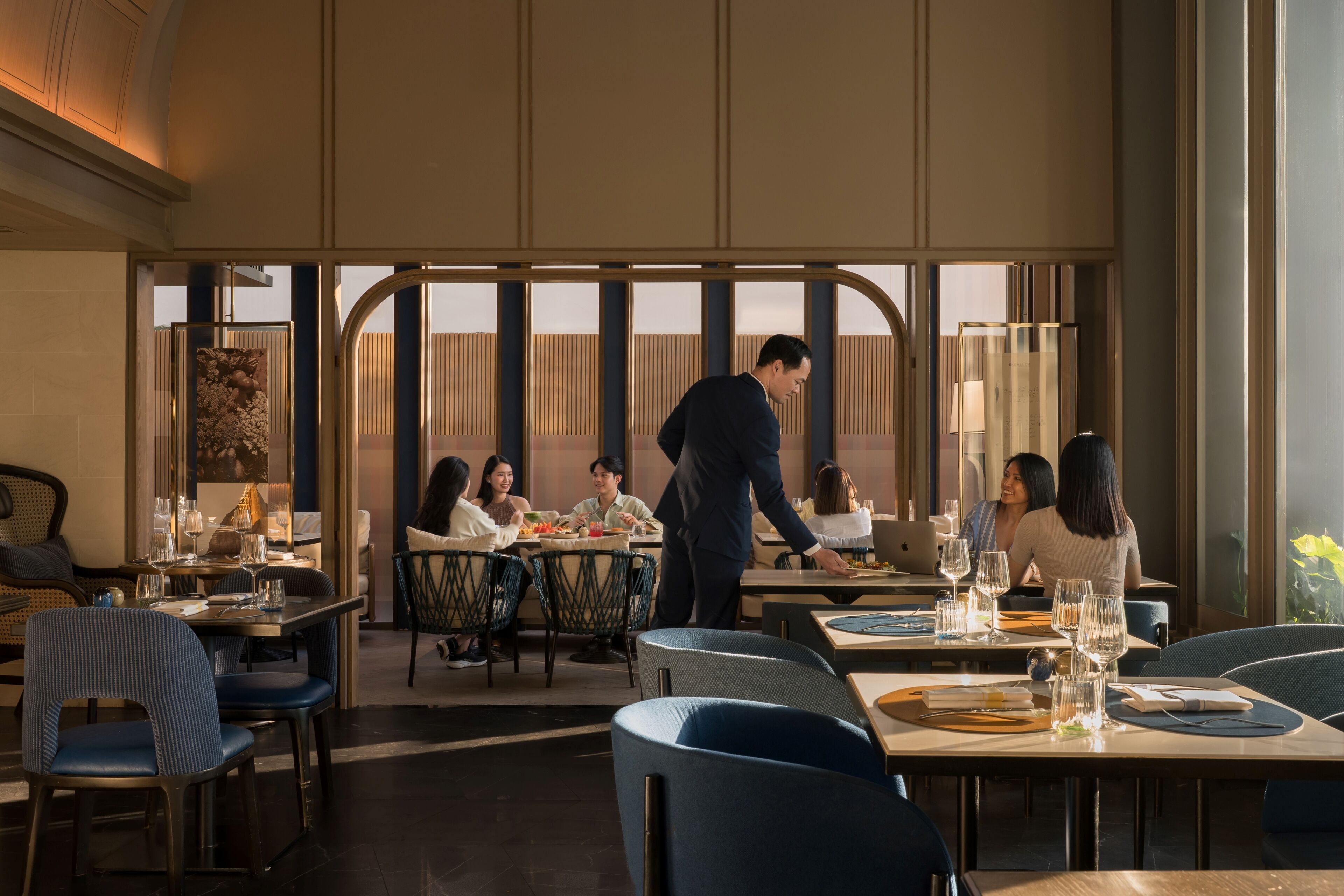 A restaurant with tables set for dining, a waiter attending to guests, and a view through large windows.