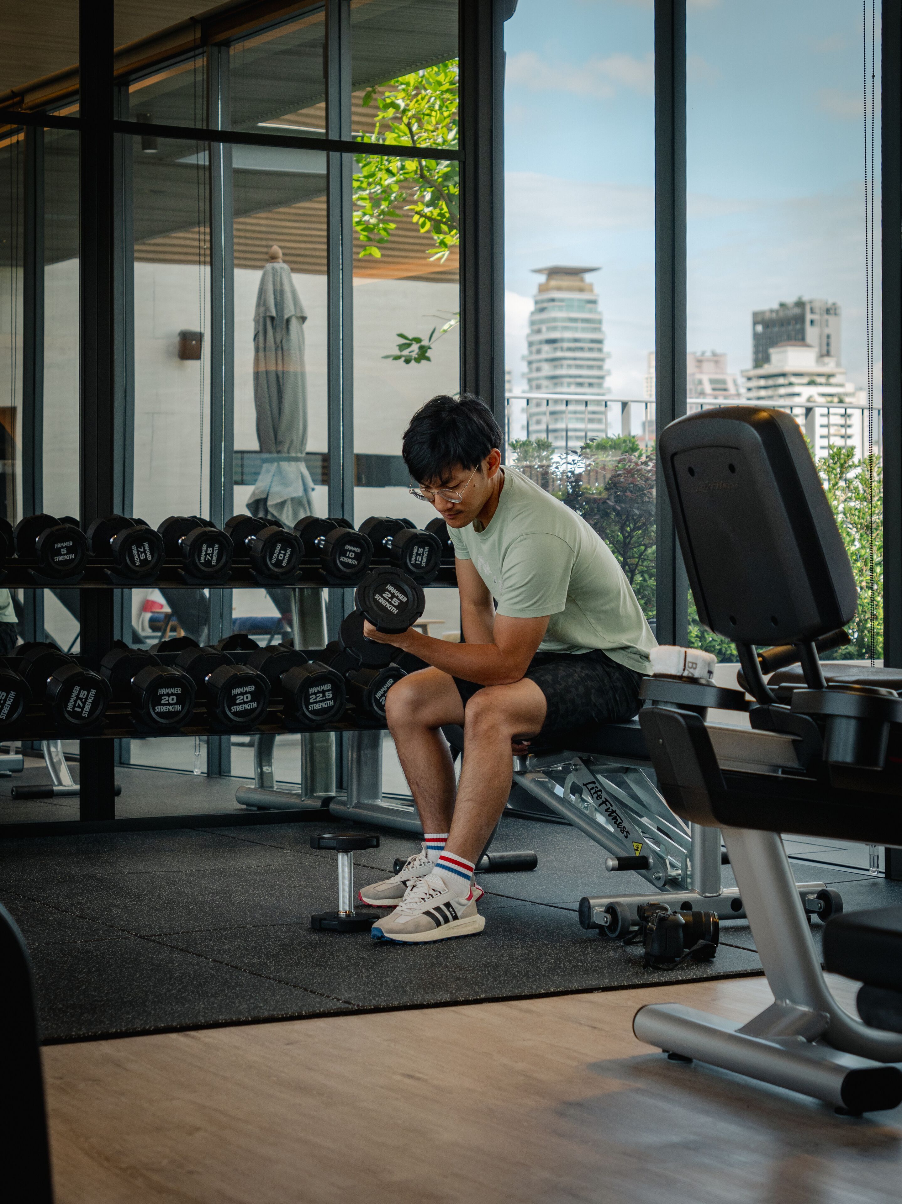 Une salle de sport équipée de matériel de musculation, d’un banc et offrant une vue sur les bâtiments à travers de grandes fenêtres.