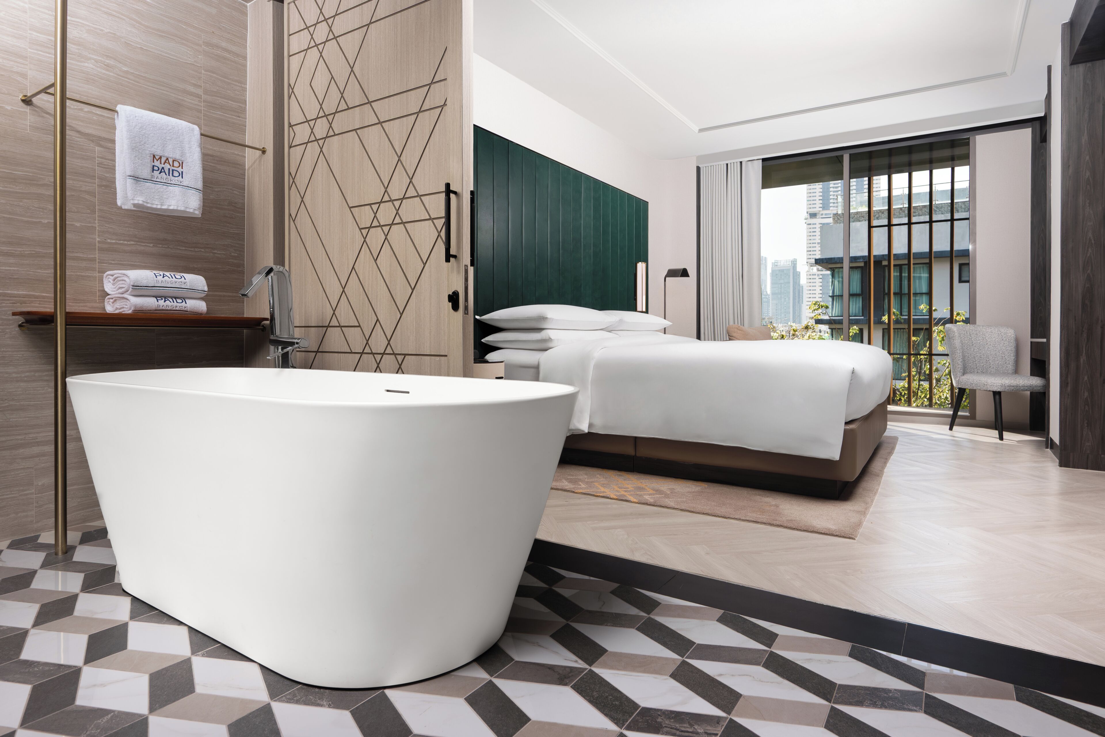 A modern hotel room with a large bathtub, a bed with white linens, a wooden wardrobe, and a view of the city through large windows.