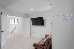 Una habitación con puerta blanca, sofá marrón, televisor montado en la pared y ventana con persianas.