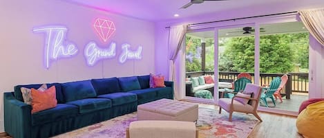 A living room with a blue sofa, a neon sign reading 'The Grand Jewel,' and a view through a glass door to an outdoor deck.