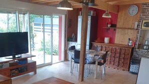 A spacious living area with a wooden coffee table, a sofa, and a TV on a wooden stand. There is a dining area with a table and chairs, and a kitchen area with a clock on the wall. - Maison "La Miloune" au Coeur de la Provence Verte, Dans Quartier Calme et Arboré (Le Val)