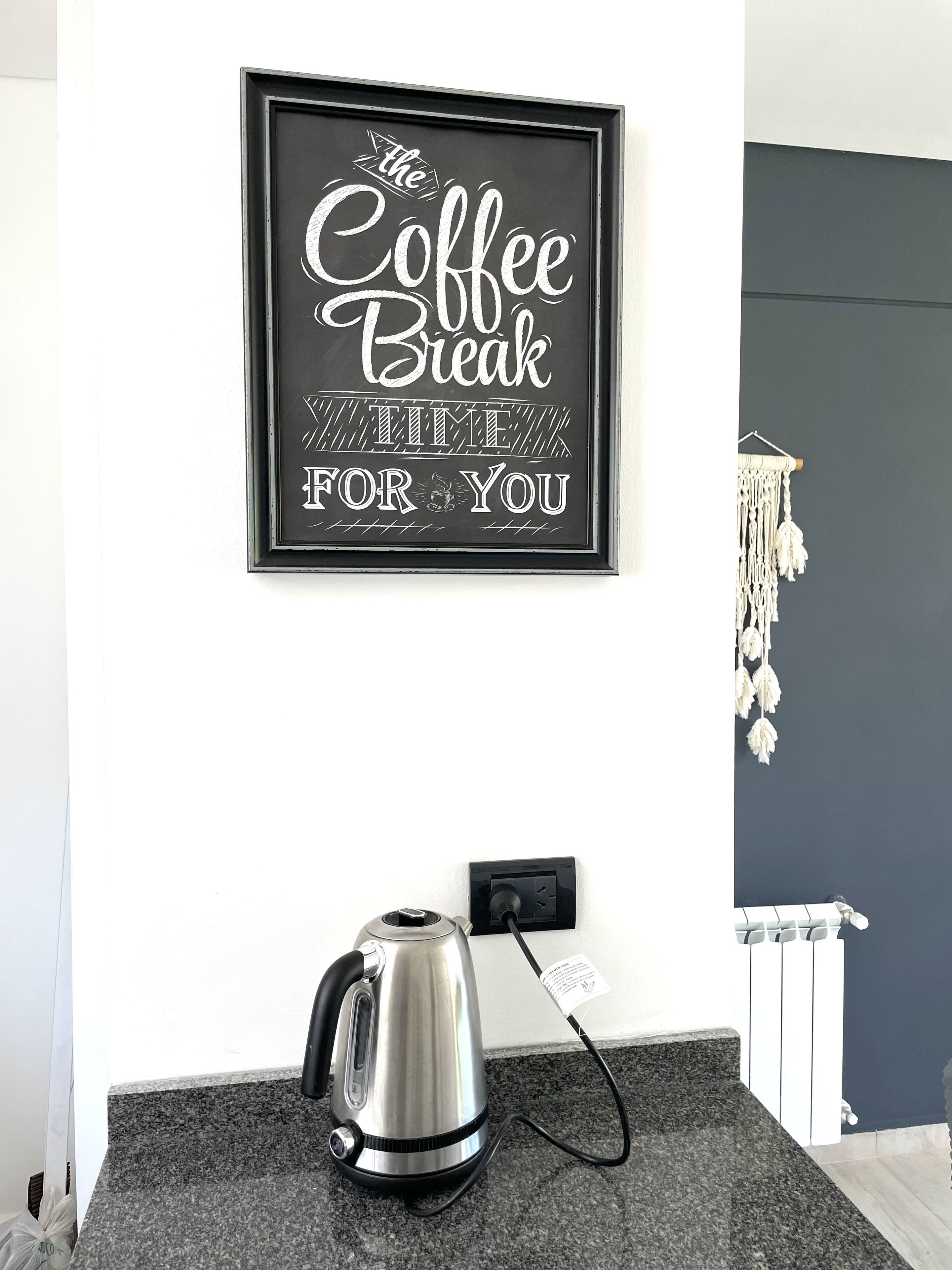 A framed sign with the text 'The Coffee Break TIME FOR YOU' on a wall, a kettle on a countertop, and a macrame wall hanging.