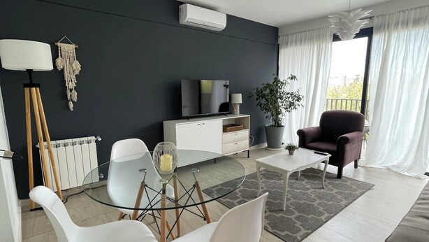 Una sala moderna con una mesa de centro de vidrio, sillas blancas, una pared de acento oscura, un televisor y una planta en maceta.