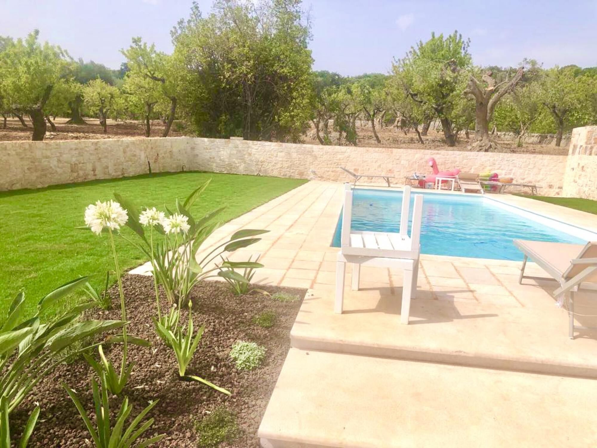 A backyard with a swimming pool, a lawn, and a stone wall.