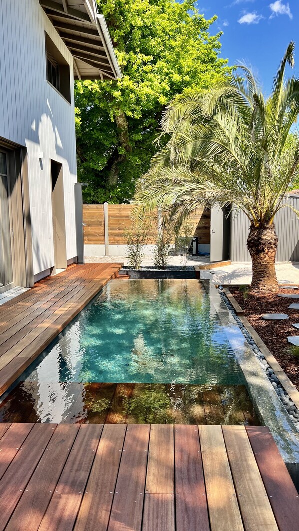 A modern outdoor pool area with clear water, wooden deck, and surrounding greenery.