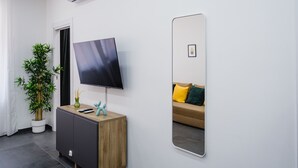 Una habitación moderna con televisión de pantalla plana, planta en maceta, mueble de madera, espejo y sofá con cojines.