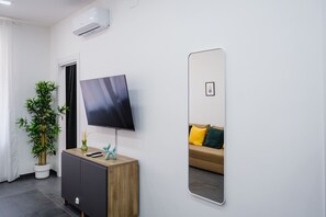 Une chambre moderne équipée d’un téléviseur à écran plat, d’une plante en pot, d’une armoire en bois, d’un miroir et d’un canapé avec des coussins.