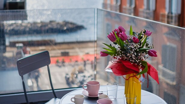 Un balcón con una mesa dispuesta para dos personas, una silla y un jarrón con flores.
