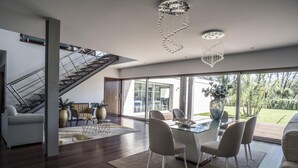 A modern living room with a glass dining table, comfortable seating, a staircase, and large windows offering a view of the outdoors.