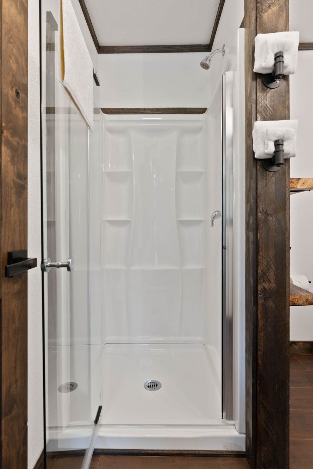 Cabina de ducha con paredes de vidrio, interior blanco, marco de puerta de madera y un perchero en la pared.