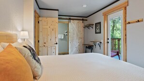A bedroom with a large bed, a wooden door, a small table, and a balcony with a view of trees. - Lake Village Lodge (Coeur d'Alene)