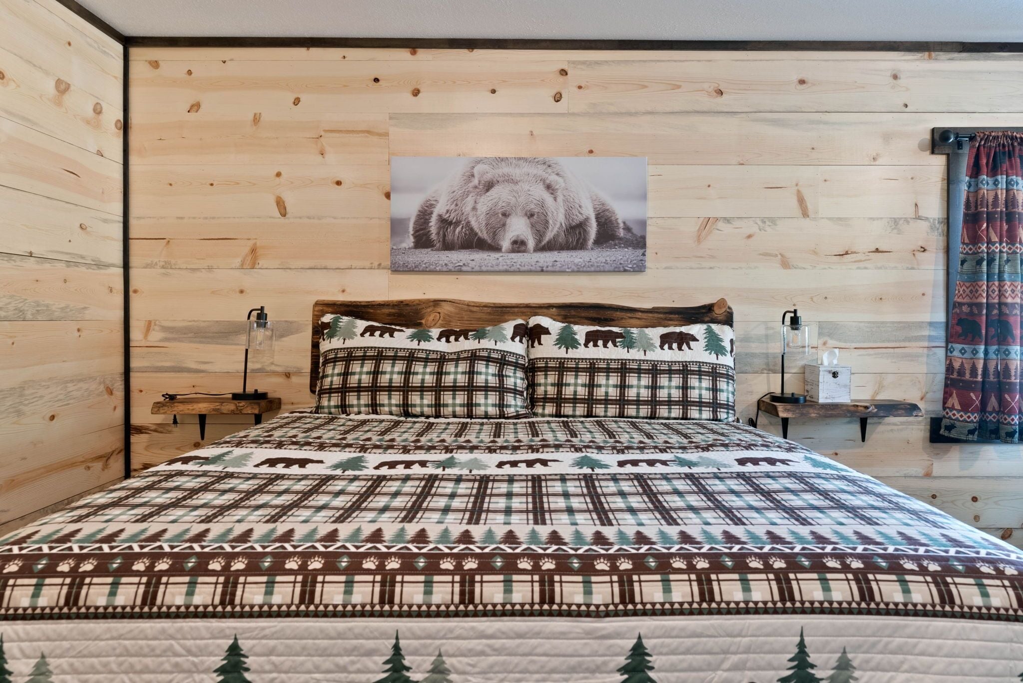 A wooden-paneled room with a bed featuring a bear-themed quilt, a mounted bear photograph on the wall, and a nightstand with a lamp.