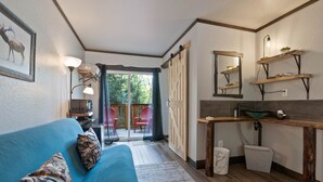 A modern living room with a blue sofa, patterned cushions, a coffee table, and a kitchen area with wooden shelves, a mirror, and a sink. - Lake Village Lodge (Coeur d'Alene)