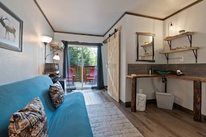 Una sala de estar moderna con un sofá azul, cojines estampados, una mesa de centro y una cocina con estanterías de madera, un espejo y un lavabo.