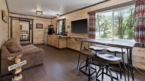 A cozy cabin interior with a wooden interior, a sofa with a moose decoration, a bar area with stools, and a large window offering a view of trees. - Lake Village Lodge (Coeur d'Alene)