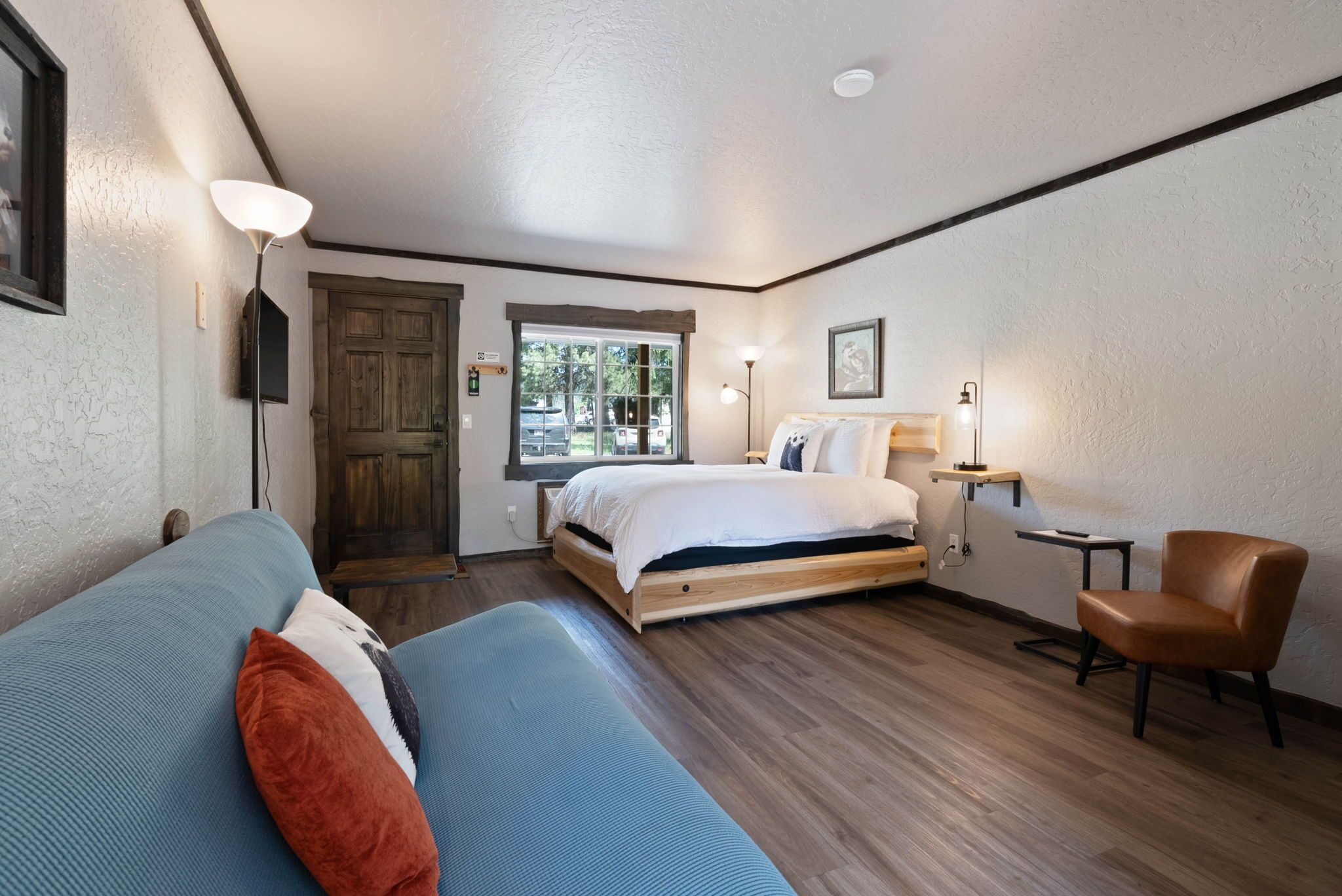 A bedroom with a bed, a blue sofa, a brown chair, a window, and a door.