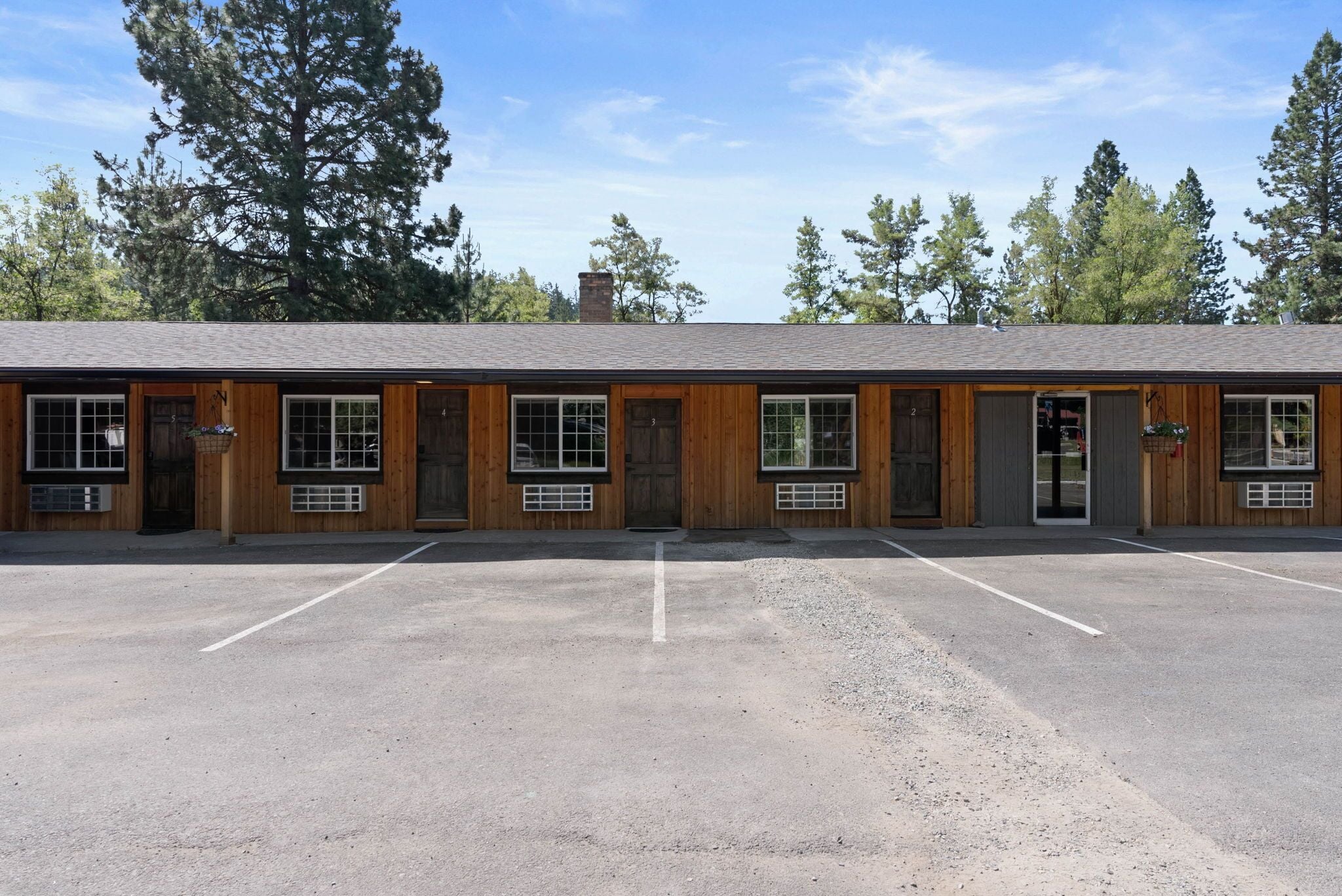 A motel with a row of rooms, each with a window and a door. There is a parking lot in front of the building.