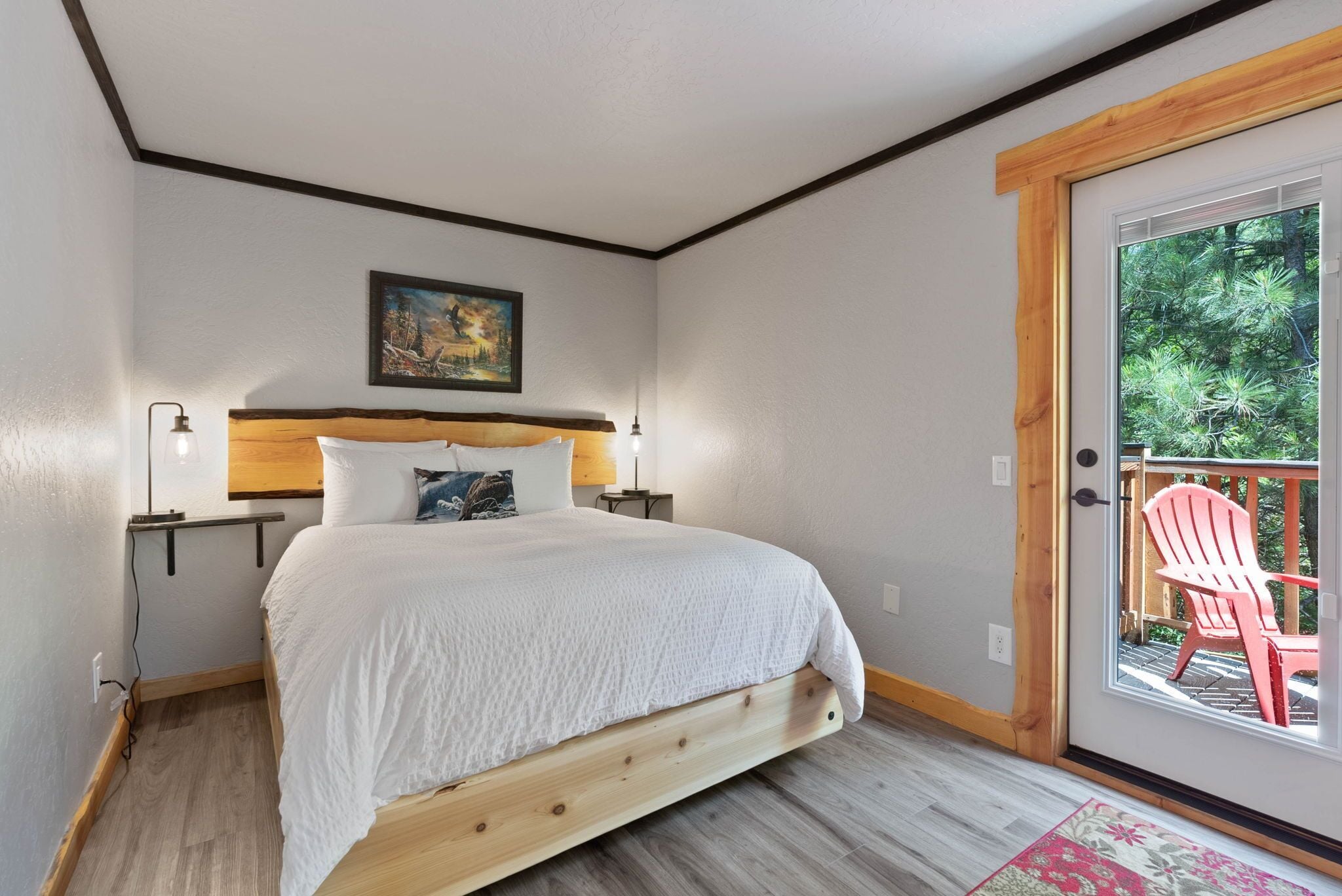 A bedroom with a wooden bed, a painting on the wall, a nightstand, and a door leading to an outdoor area with a chair.