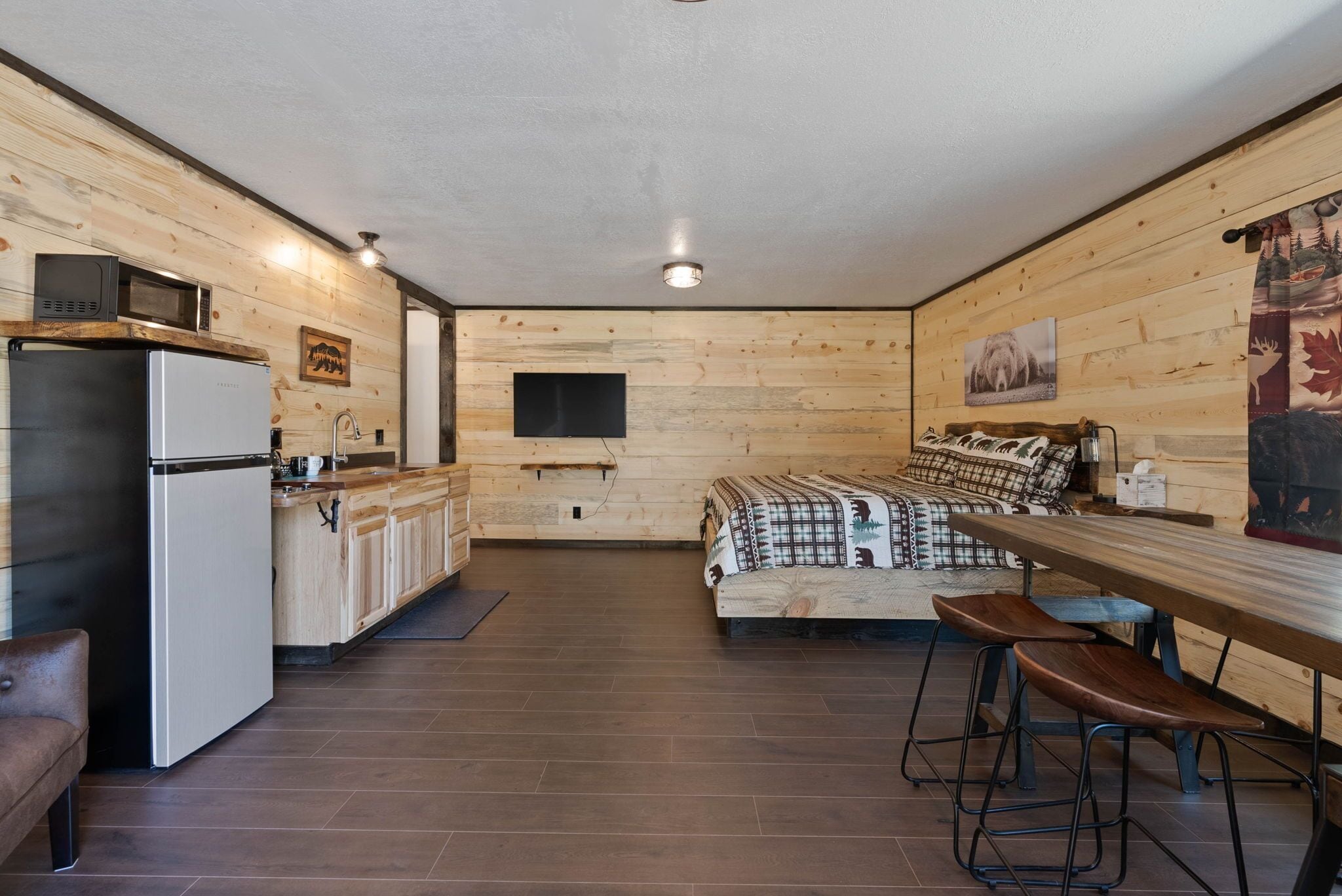 A compact kitchen with a refrigerator, microwave, and wooden cabinets. There is a bed with a patterned blanket and two wooden stools.