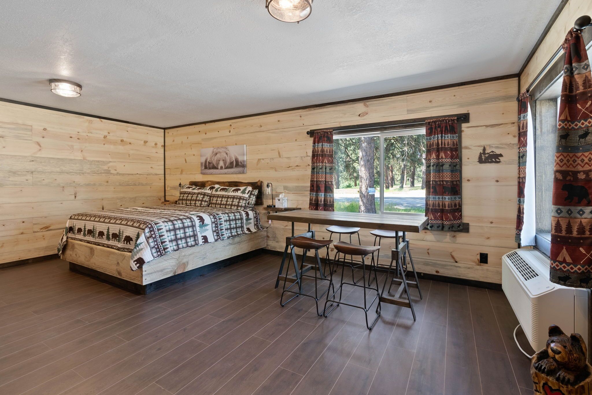 Ein Zimmer mit einem Bett, einem Tisch mit Hocker, einem Fenster mit Vorhängen und einer Bär-Dekoration.