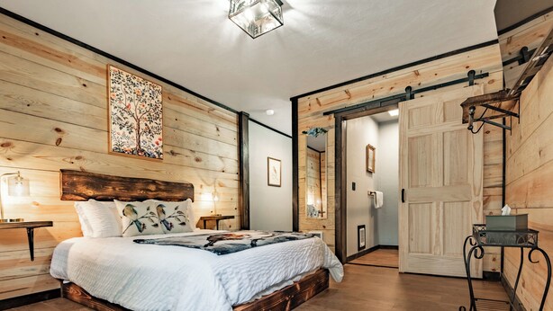 A bedroom with a wooden headboard, a large bed with a floral bedspread, a nightstand with a lamp, and a sliding door leading to another room. - Lake Village Lodge (Coeur d'Alene)