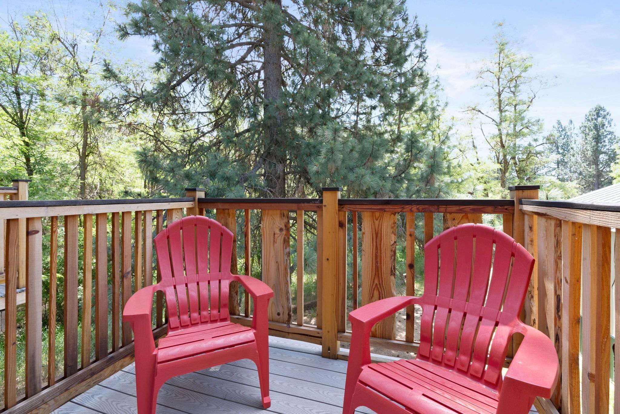 A wooden deck with two red Adirondack chairs facing each other, surrounded by trees.