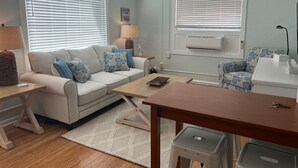 A living room with a sofa, coffee table, and a small table with a lamp. There is a ceiling fan, a wall-mounted air conditioner, and a window with blinds. - Tropical Hideaway in Downtown Flagler Beach (Flagler Beach)