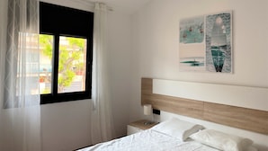 Ein Schlafzimmer mit einem Bett, einem Nachttisch, einer Lampe und einem Fenster mit transparenten Vorhängen.